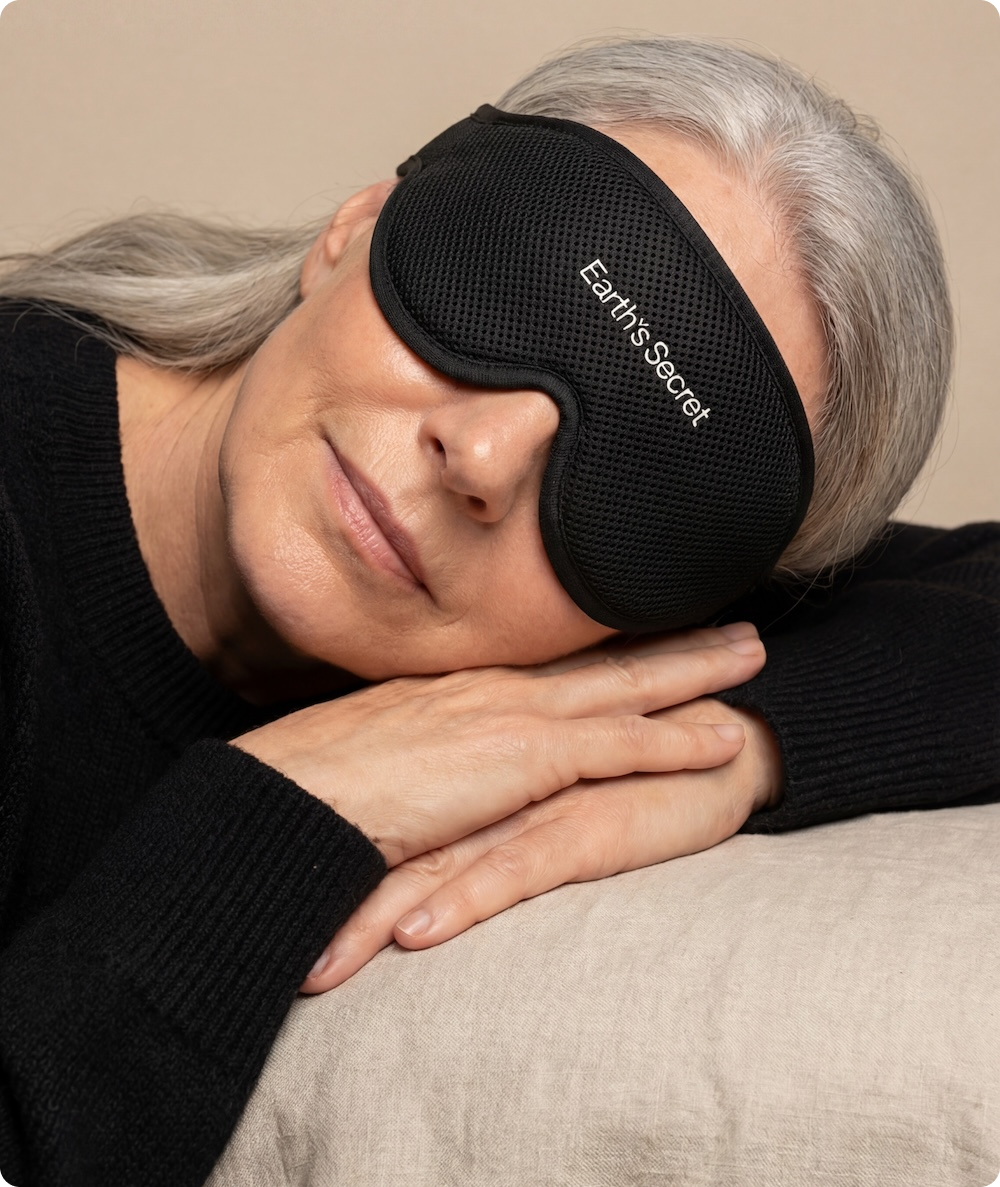 Woman wearing a black sleep mask with 'Earth's Secret' branding, resting her head on a beige surface.