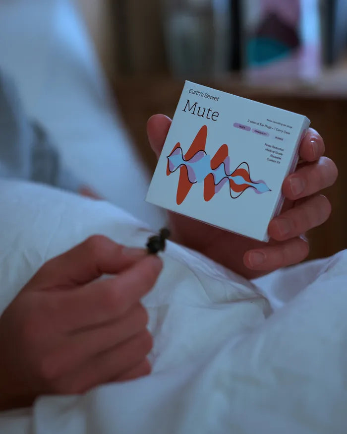 Hand holding a Mute ear plug and the Mute box in bed
